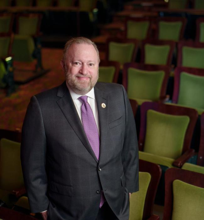 Headshot of Jay Bernhardt, Ph.D., MPH, former Dean of Moody College of Communication, current President of Emerson College, and founding director of the Center for Health Communiation at The University of Texas at Austin. A white man with a graying beard in a dark gray suit and purple tie.