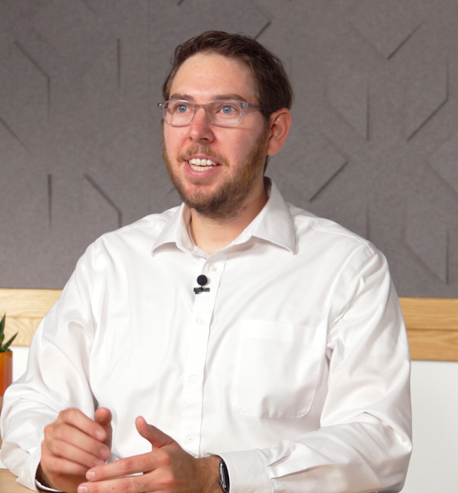 A photo of Dr. Spencer J. Fox, a white man with short, brown hair wearing glasses and a white button-down shirt sitting at a desk and talking to a camera
