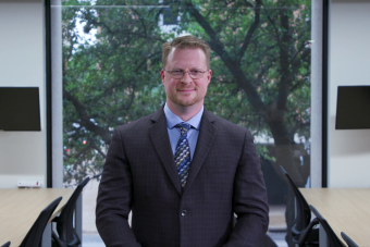 Snapshot of the Health Communication with the Deaf and Hard of Hearing Community Health Communication Training Series course taught by Dr. Chris Moreland. A white man with short blonde hair and glasses standing in front of a window in a dark suit and tie looking at the camera with a closed-mouth smile.