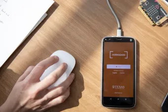 A top view of a computer desk with an iPhone plugged in to a charger next to a hand using a computer mouse. On the phone is the login screen for the Hornsense app from The University of Texas at Austin.
