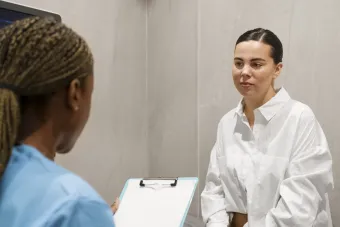 A photo of a young woman wearing a white blouse sitting in a doctor's office speaking to a nurse.
