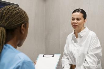 A photo of a young woman wearing a white blouse sitting in a doctor's office speaking to a nurse.