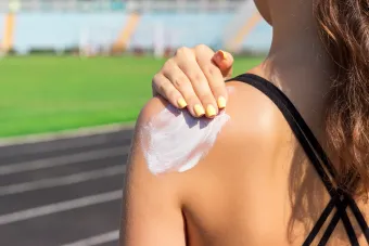 A person with yellow-painted fingernails applies sunscreen to their shoulder. The background features a blurred running track and green field, indicating an outdoor setting.