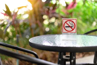 A round glass-top table with black metal edges and chairs set outdoors in a garden area. A small sign with a no-smoking symbol is placed on the table, indicating smoking is not allowed. The background shows blurred greenery with sunlight filtering through the leaves.