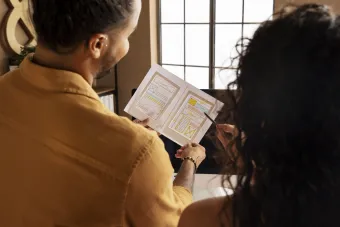 A man and a woman stand together in a well-lit room with a large window in the background. The man holds an open booklet with colorful diagrams and text, while the woman points at it with a pen.