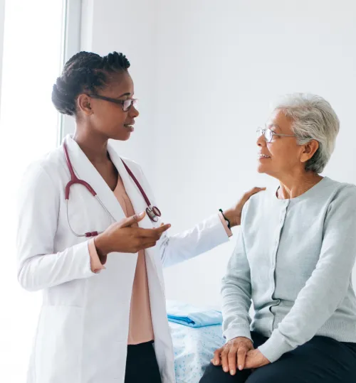 A young Black female doctor wearing a white coat and stethoscope speaking to an elderly female patient.