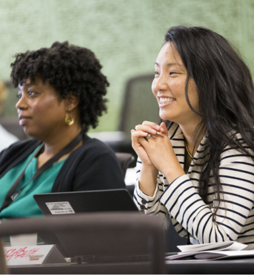 Two attendees of the Health Communication Leadership Institute - a Black woman with short curly hair in a green blouse and black cardigan sitting next to an Asian woman with long dark hair wearing a black and white striped shirt. Both are looking off to the side at someone speaking.