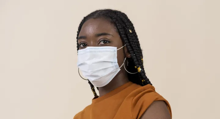 A young Black woman with long, braided hair wearing a sleeveless orange top and a face mask.