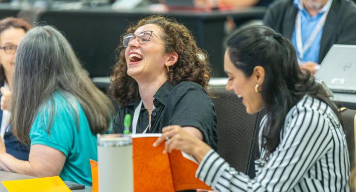 Two women sitting in a classroom at the 2025 Health Communication Leadership Institute in Austin, TX. One woman has long, curly, brown hair and is wearing glasses and a short-sleeve button down shirt. The other woman has long, black hair and is wearing a striped, long-sleeve button-down shirt.